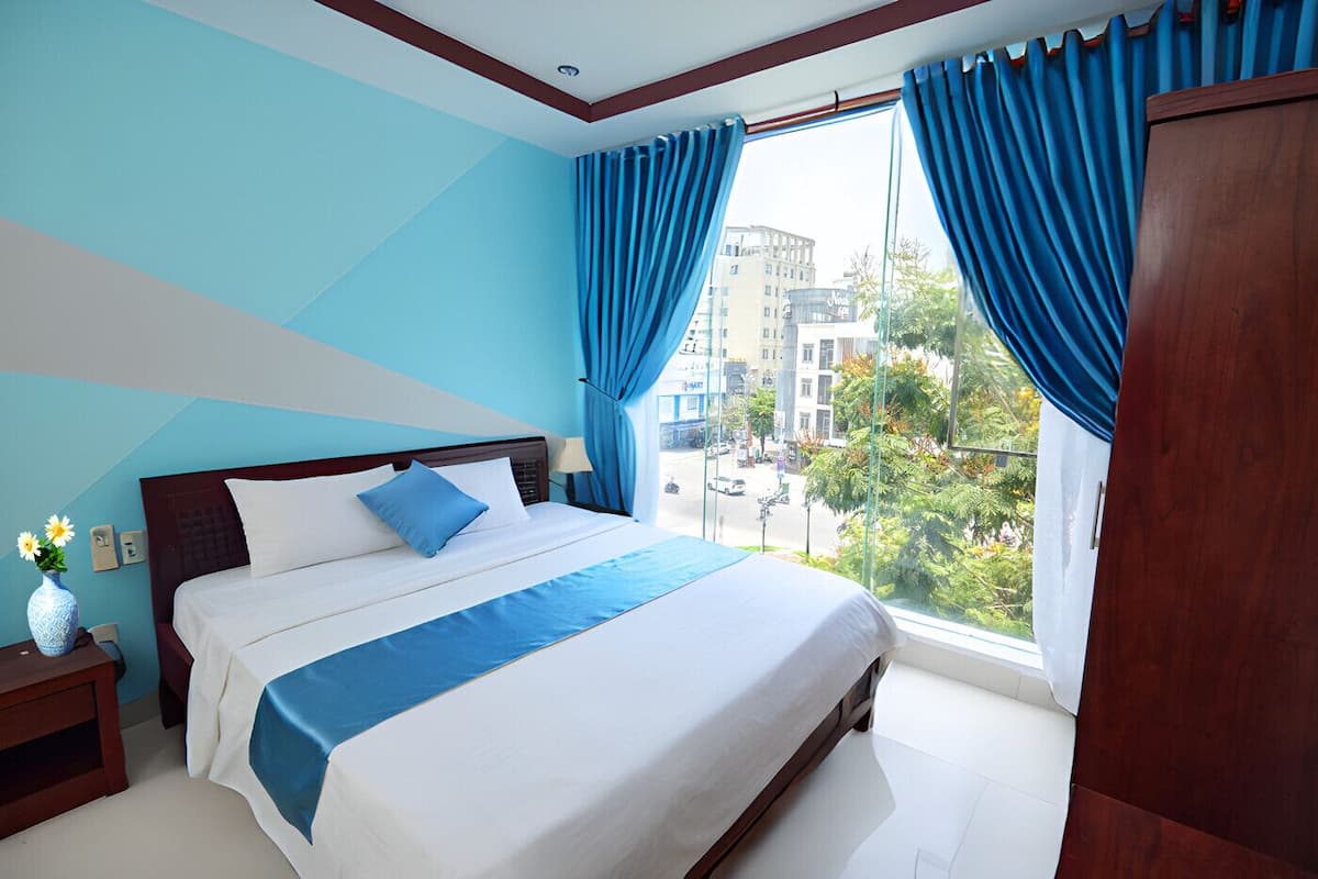 A modern hotel room with a large bed, a nightstand, a vase with flowers, and a view of the city through large windows.
