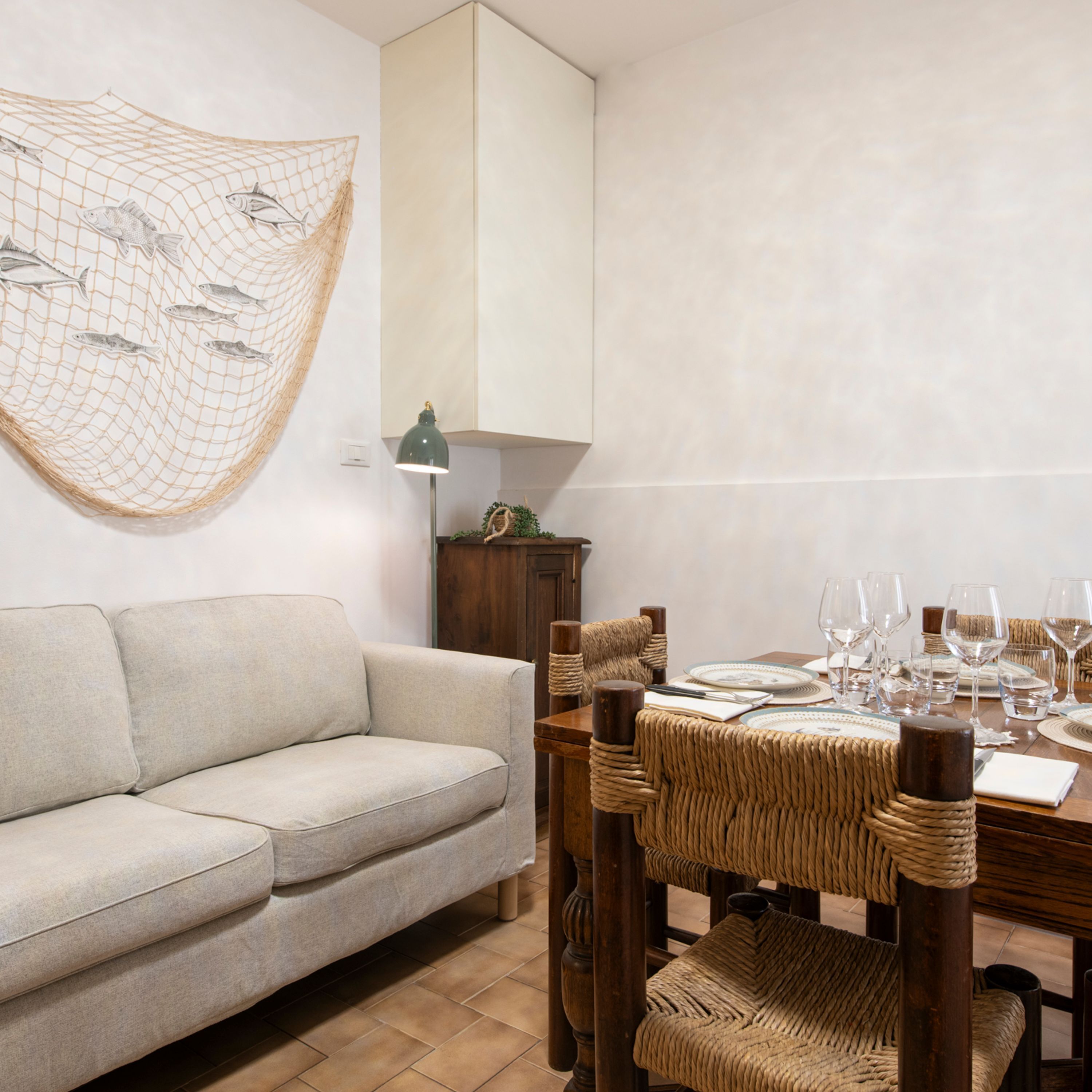 A dining area with a wooden table set for four, a woven chair, a sofa, and a wall-mounted fish net decoration.
