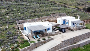 Un edificio blanco con detalles azules, muros de piedra y un área pavimentada con sillones de descanso.