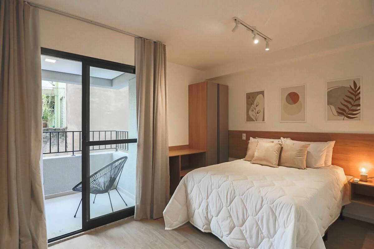A hotel room with a bed, a nightstand, a chair, a balcony with a view, and artwork on the wall.