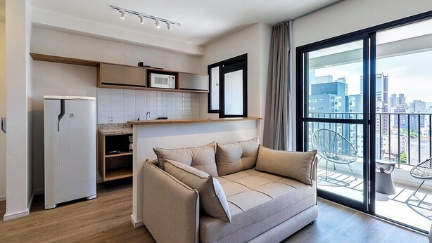 A modern apartment with a kitchenette, a sofa, and a balcony with a city view. - 360 Pulse Higienópolis  (São Paulo)