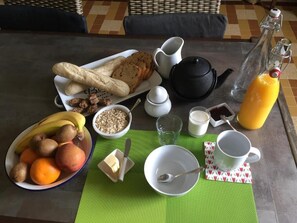 A breakfast table with bread, fruits, a teapot, a milk jug, and a bottle of orange juice. - L'intemporelle (Ozourt)