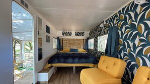 A compact RV bedroom with a bed, two yellow chairs, a small table, and a window with a view of the outdoors. - L'intemporelle (Ozourt)