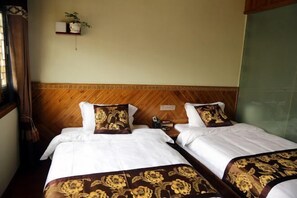 A hotel room with two beds, a wooden headboard, a small shelf with a plant, and a window with curtains.