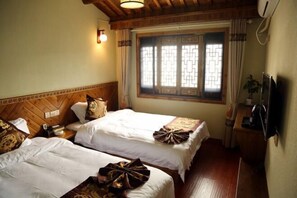 A hotel room with two beds, a wooden headboard, a window with decorative glass, a bedside table with a lamp, and a television on the wall.