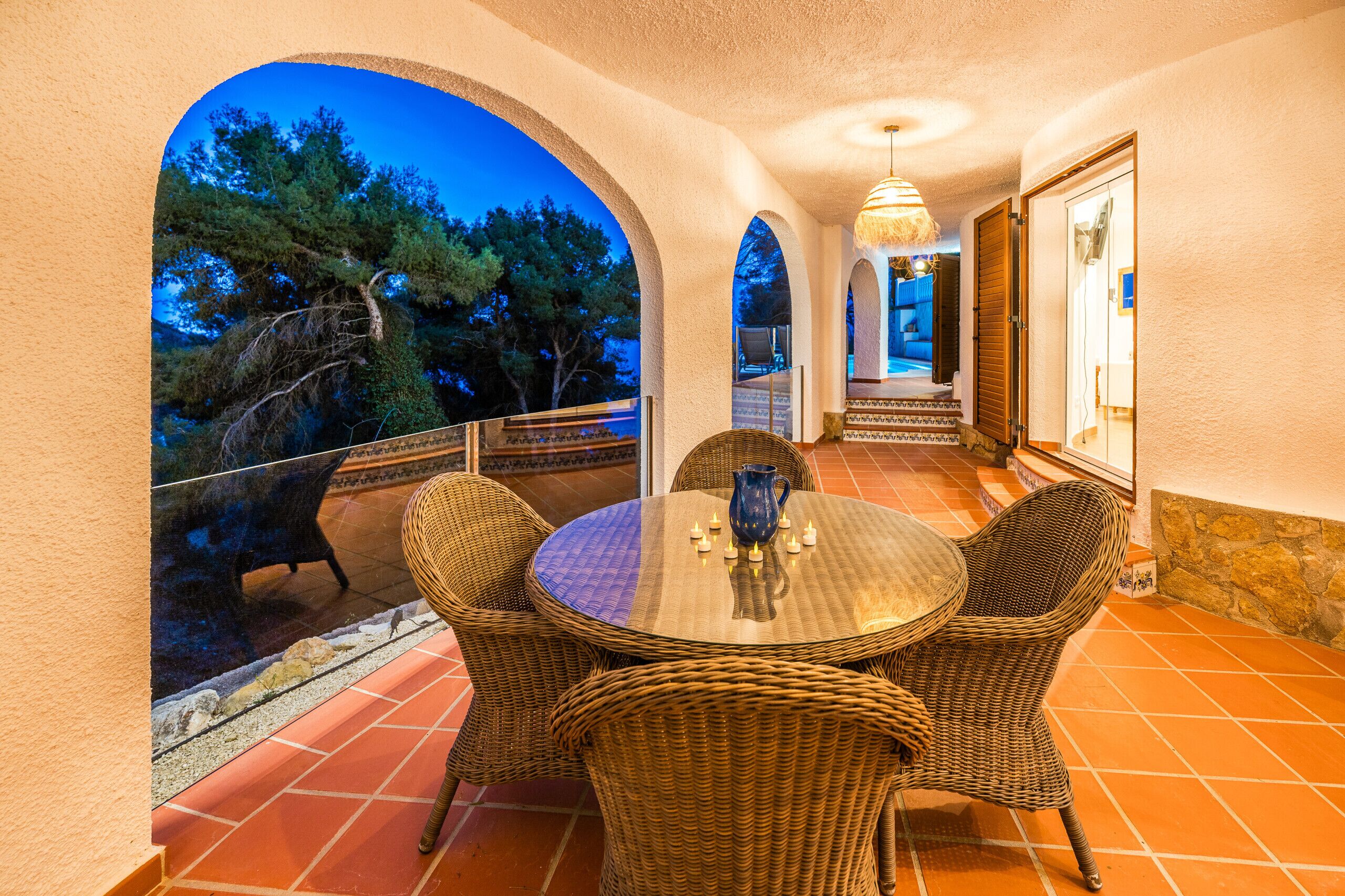 Un patio con mobiliario de mimbre y una mesa, con vistas panorámicas a árboles y al atardecer.