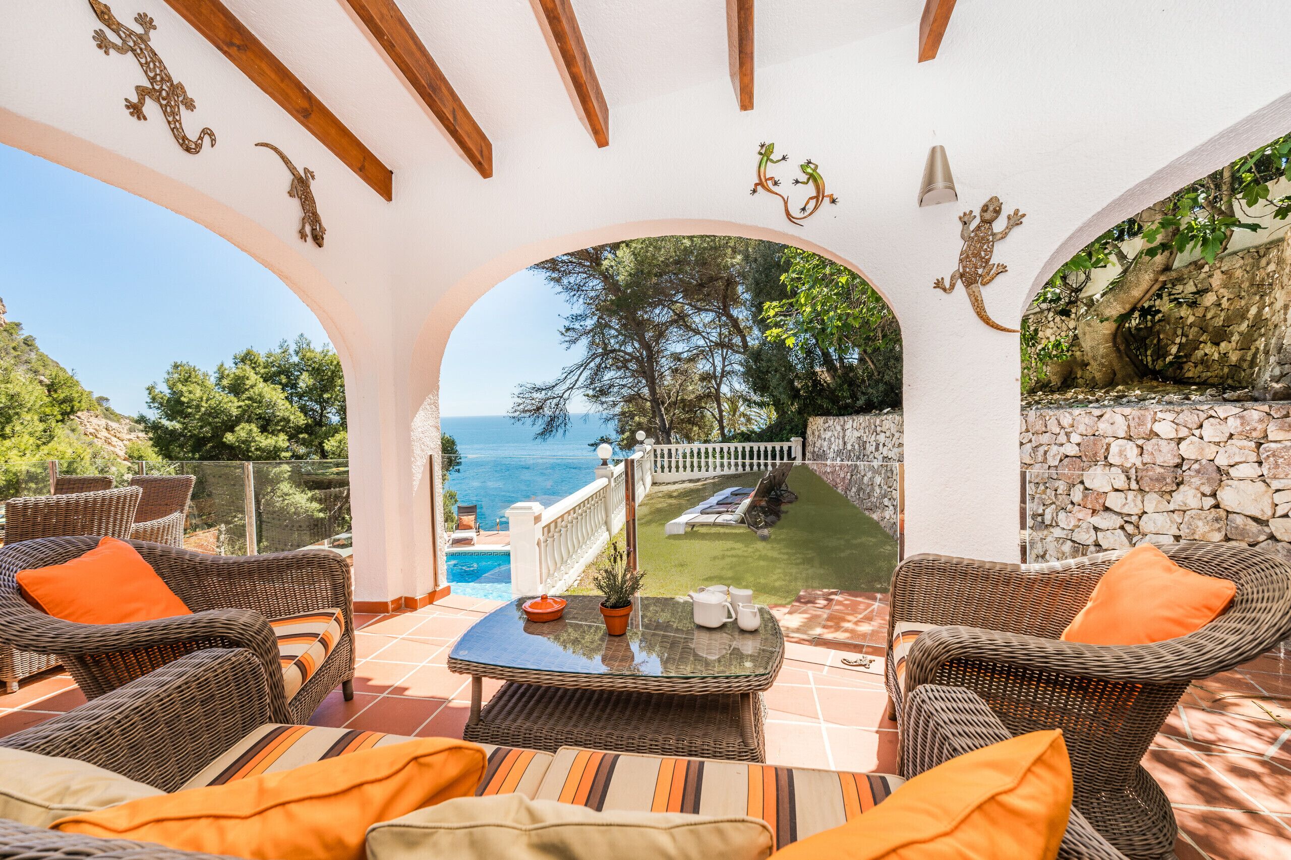 Eine überdachte Terrasse mit Korbmöbeln, Blick auf das Meer und einer Steinmauer.