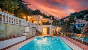 A poolside view of a Mediterranean-style villa with a stone wall, a balcony with chairs, and a view of the surrounding hills.