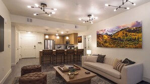 A modern living room with a sofa, ottomans, and a coffee table. There is a kitchen area with a refrigerator and a dining area with chairs. - Luxury Downtown 2 Bd 2 Ba Suite - Rooftop Terrace, Steps to Delta Center & More! (Salt Lake City)
