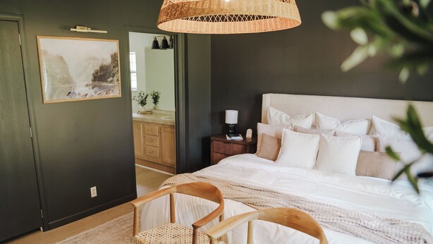 A bedroom with a large bed, two wooden chairs, a woven rug, a bedside table with a lamp, and a framed picture on the wall.