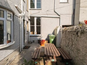Un espacio exterior estrecho con una mesa de madera y bancos, un bote de basura verde y una ventana.