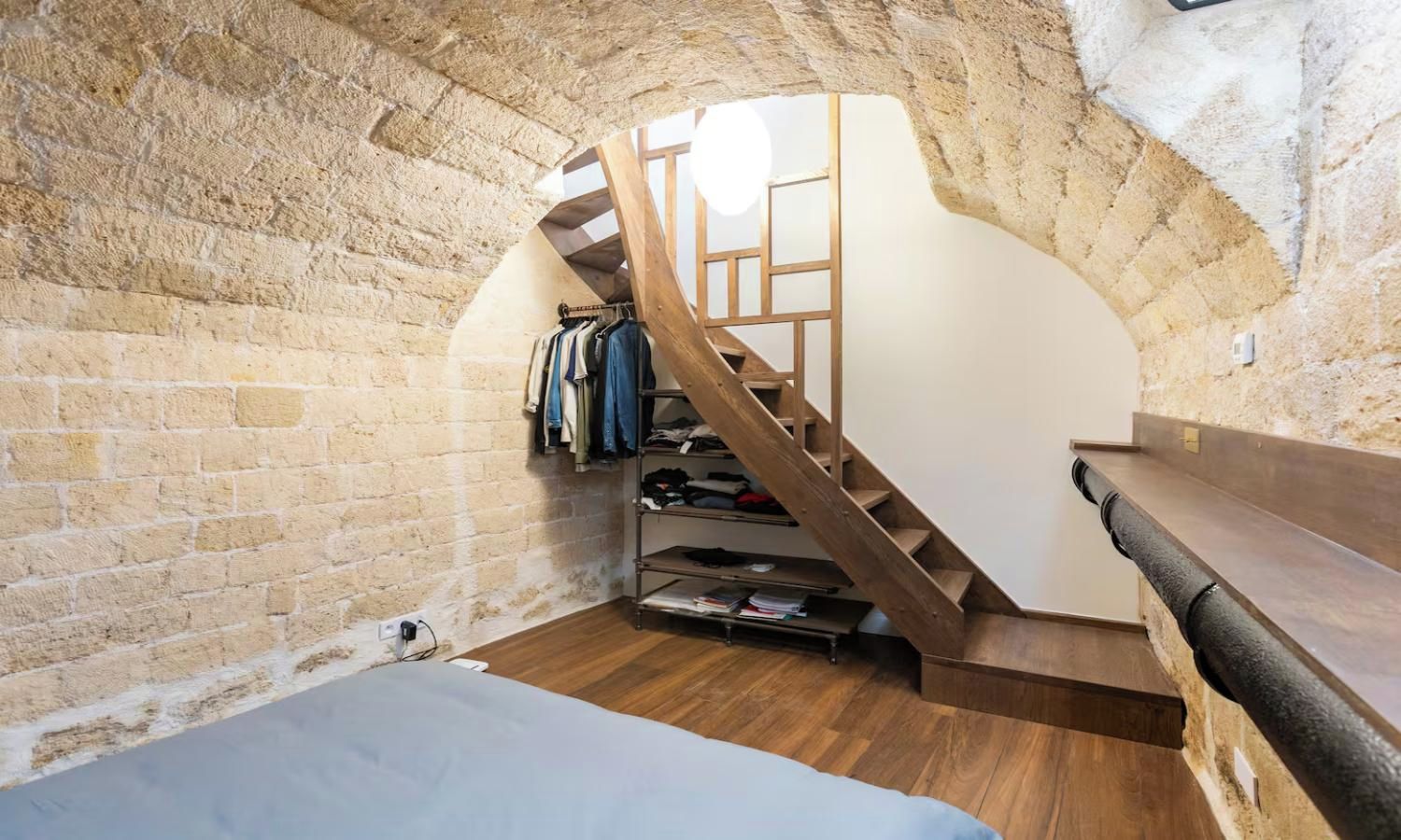 A room with a wooden staircase, a bed, a clothing rack, and a small shelf.