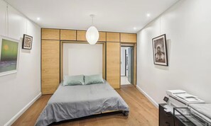 A modern bedroom with a bed, a desk with a printer, and a door leading to another room.