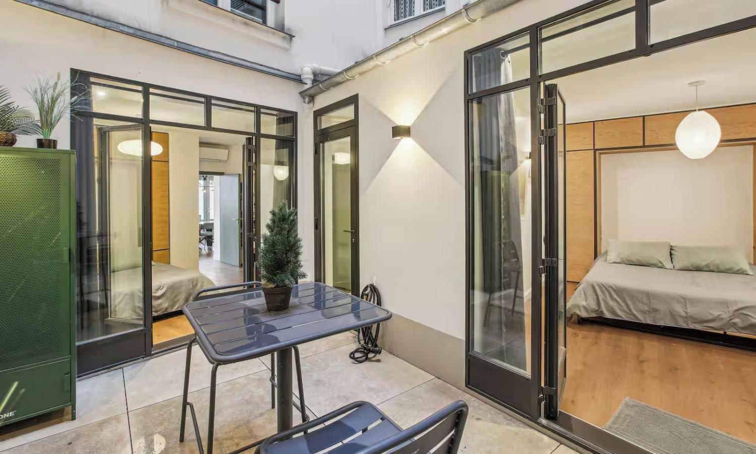 Un intérieur moderne avec une porte vitrée donnant sur un balcon, une petite table avec un pot de fleurs et un lit doté d’une tête de lit en bois.