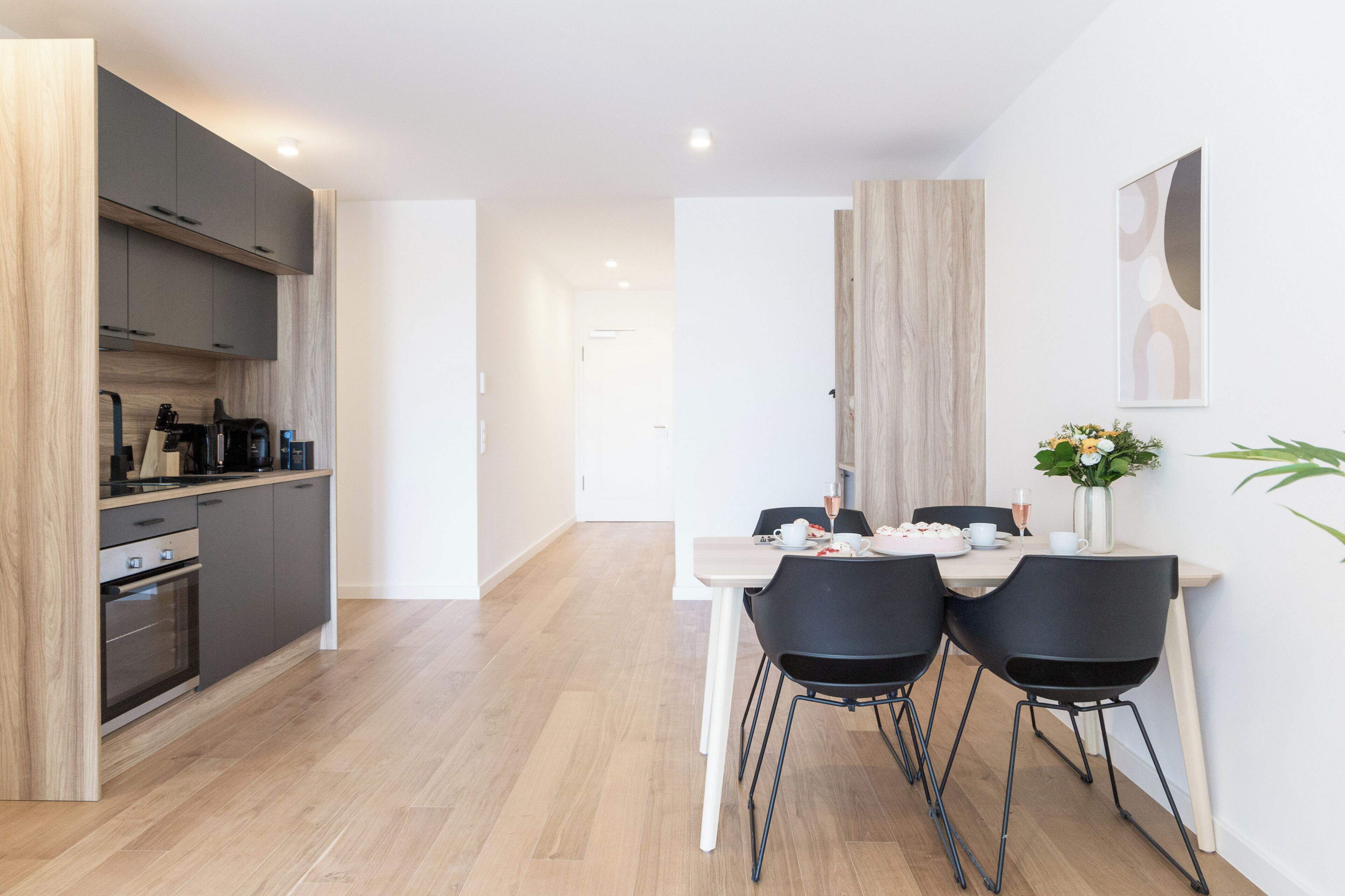 A modern kitchen with dark cabinets and a wooden countertop, a dining area with a white table and black chairs, and a wooden accent wall.