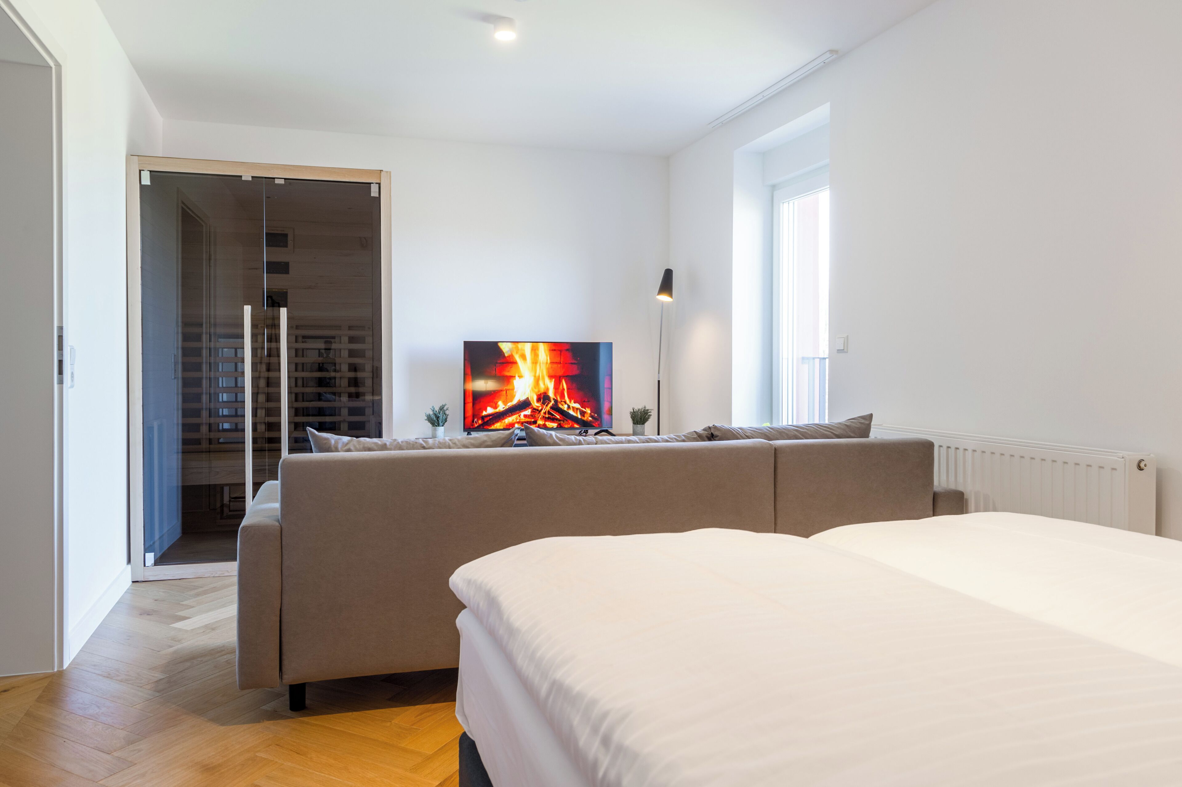 Een moderne hotelkamer met een groot bed, een bank, een televisie waarop een haard te zien is, en uitzicht op een houten vloer en een deur naar een balkon.