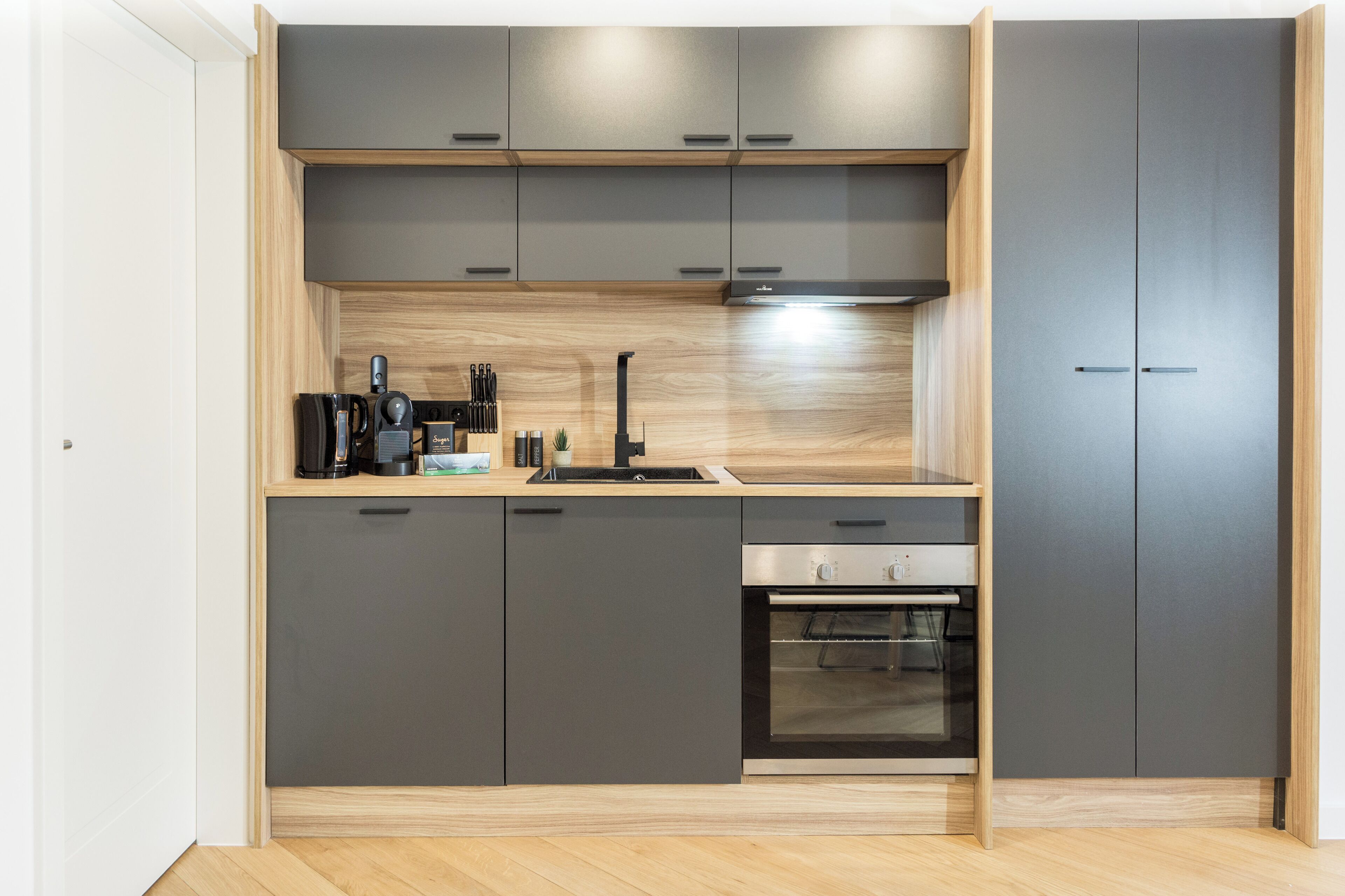 A modern kitchen with grey cabinets, a wooden countertop, a built-in oven, and a coffee machine.