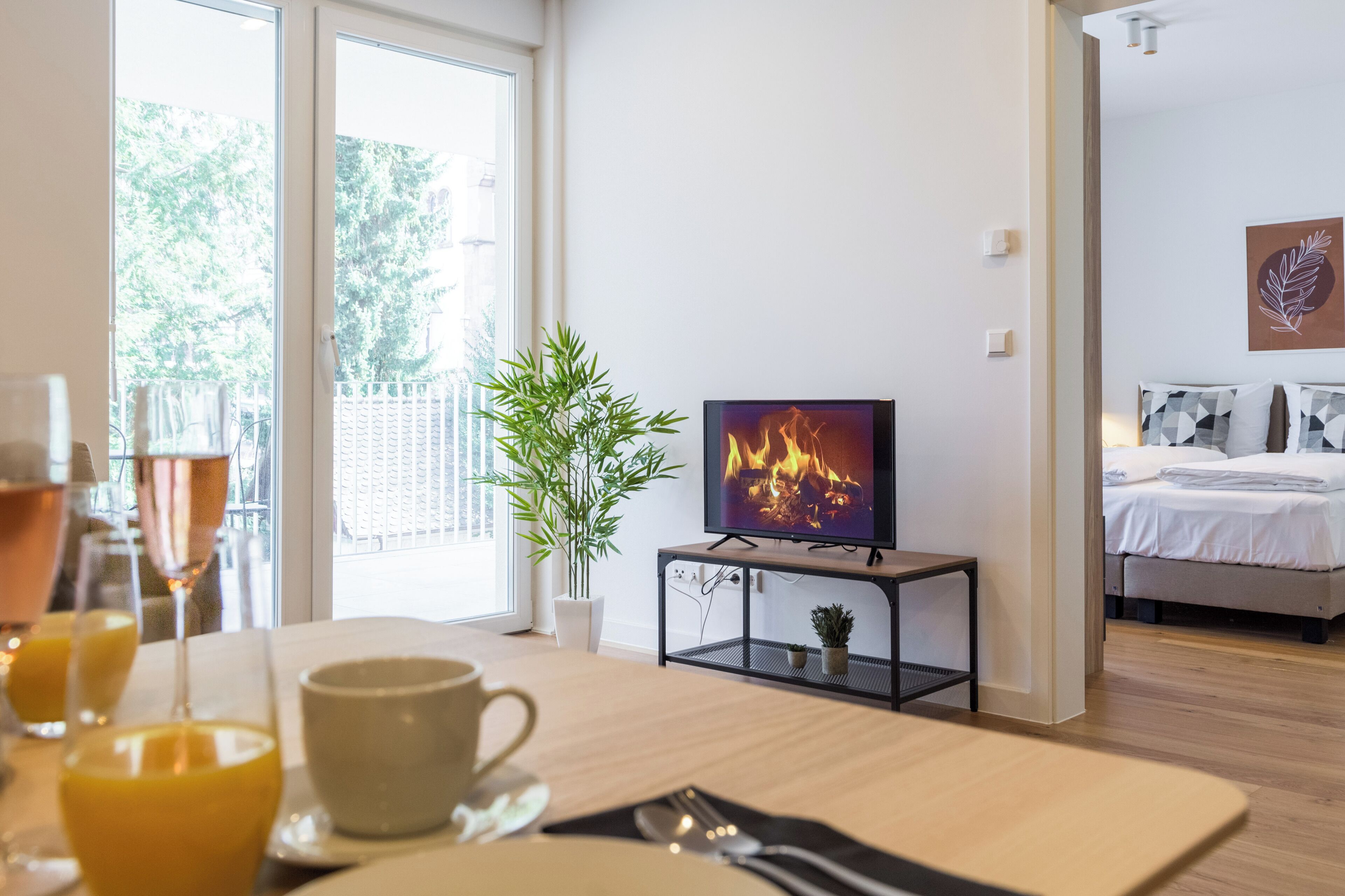 Ein modernes Hotelzimmer mit einem Bett, einem Fernseher, der einen Kamin zeigt, einem Essbereich mit Tisch für zwei Personen und einem großen Fenster mit Blick auf Bäume.