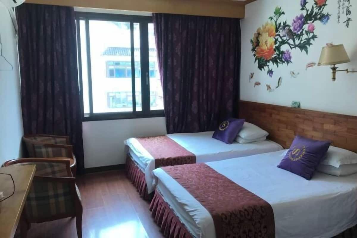 A hotel room with two beds, a wooden desk, a chair, a window with curtains, and a wall with a floral mural.
