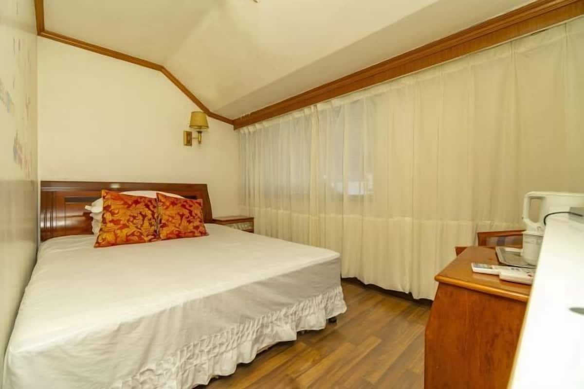 A hotel room with a bed, a wooden nightstand, a desk with a chair, and a window with curtains.