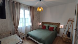 Et soveværelse med seng, sengeborde med lamper, en træsenggavl, en hængende lampe, en træstol og et vindue med gardiner.