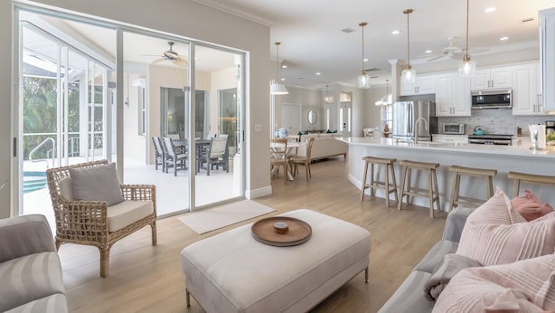 Un amplio salón con un ventanal grande, una silla de mimbre, un banco acolchado y una cocina con armarios blancos y una zona de bar.