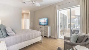 Un dormitorio con cama, televisor, ventilador de techo, puerta corrediza de vidrio a un balcón y zona de comedor visible a través de la puerta.