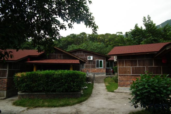 A rustic cabin-style building with a brick exterior and a green door, surrounded by lush greenery and a concrete pathway.