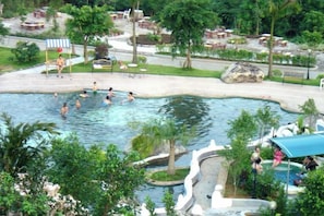 Une piscine où se baignent des personnes, entourée de verdure et bordée d’un petit bâtiment.