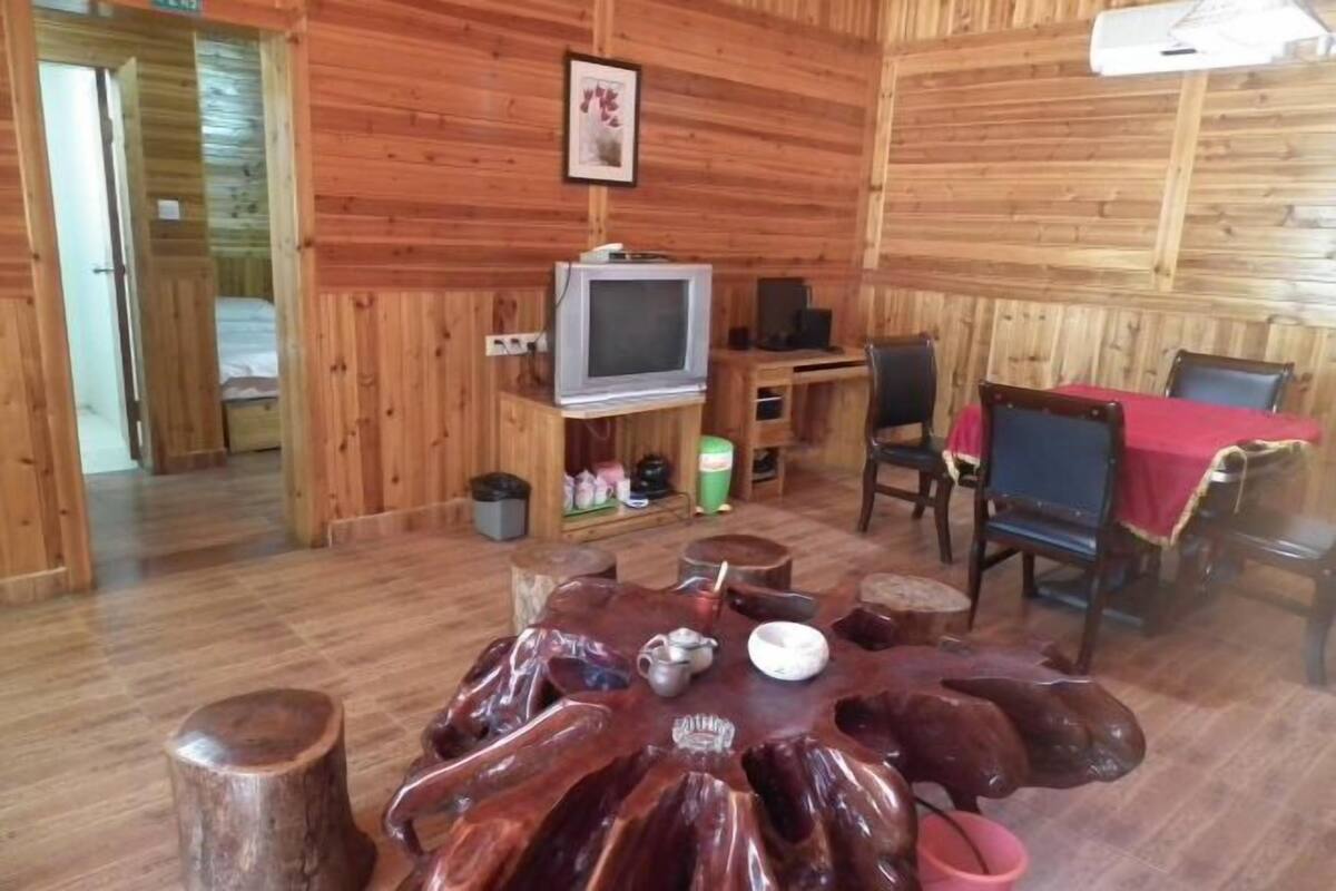 Interior de una cabaña de madera con una mesa redonda, sillas, un televisor y una cama al fondo.