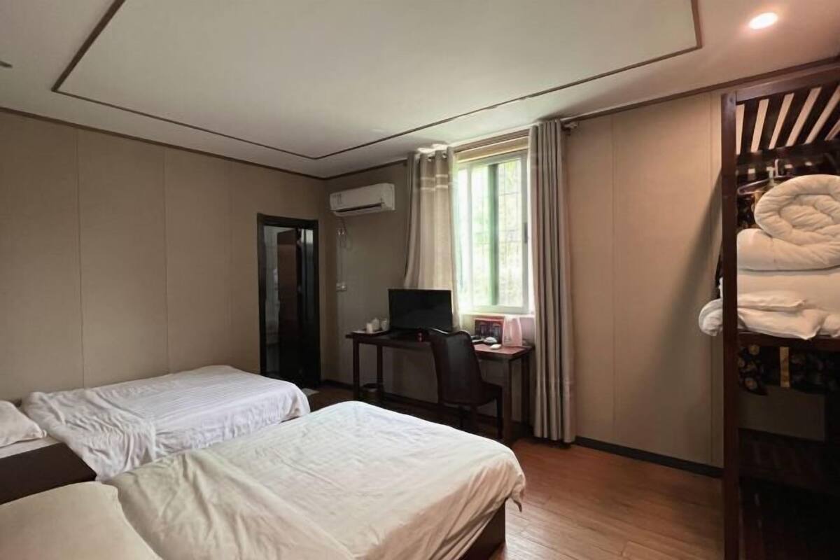 Habitación de hotel con dos camas, un escritorio con computadora, un equipo de aire acondicionado, una ventana con cortinas y una litera con toallas.