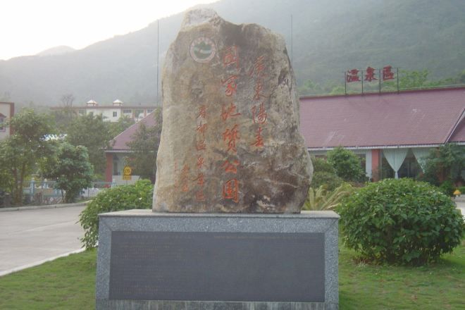 A stone monument with inscriptions, a paved road, and a building with a red roof in the background.