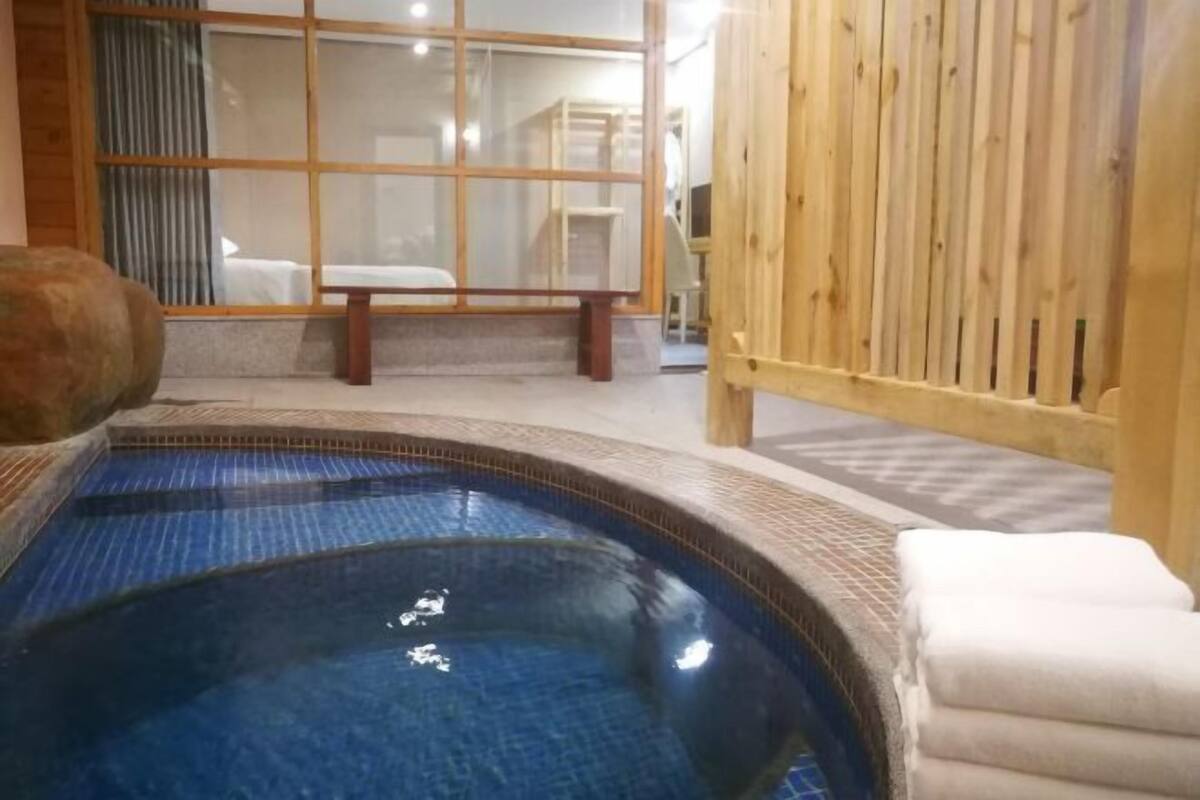 Zona de hidromasaje con valla de madera, un banco y vista a una habitación con cama y ventana.