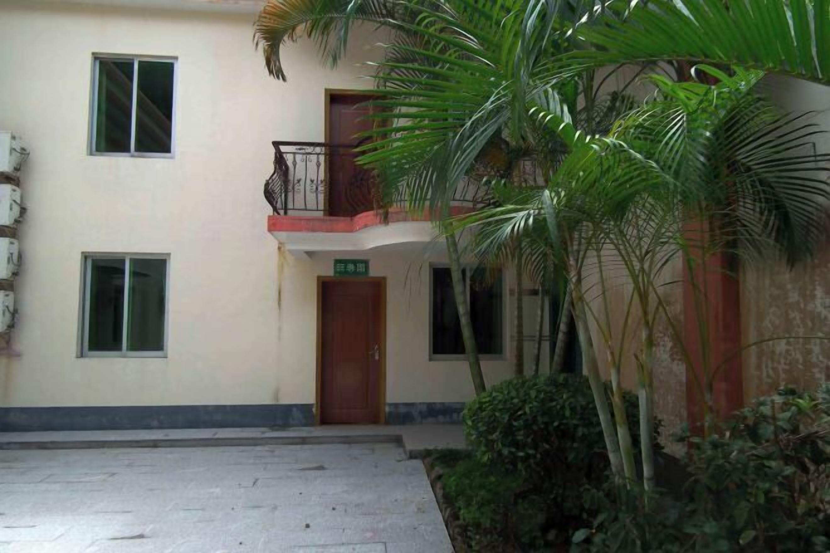 A building with a ground floor entrance, a balcony with a metal railing, and a small garden area with palm trees.