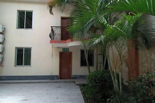 Un edificio con entrada a nivel de calle, un balcón con barandilla metálica y un pequeño jardín con palmeras.