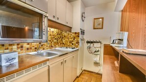 A compact kitchen with a microwave, stove, and sink. There is a wooden cabinet and a tiled backsplash. A coffee maker is on the counter.