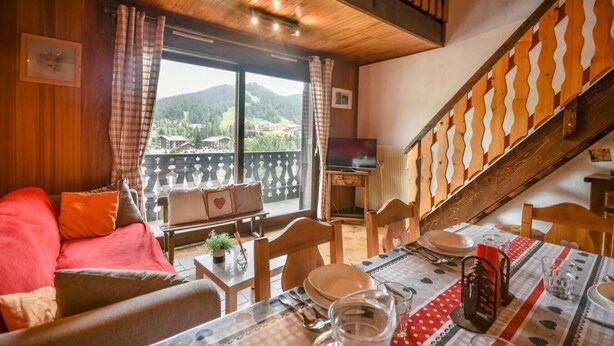 A cozy room with a wooden ceiling, a balcony with a view of the mountains, a dining table set with plates and glasses, and a staircase with a wooden railing.