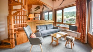 A cozy room with a wooden staircase, a sofa, a coffee table, and a view of a village through large windows.
