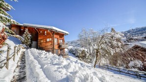 A wooden cabin with a balcony, surrounded by snow-covered trees and a clear blue sky.
