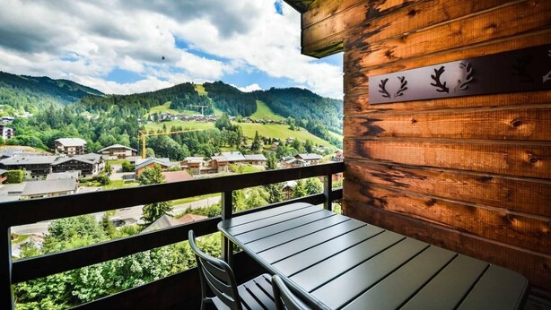 A wooden balcony with a table and chairs, overlooking a village and mountains.