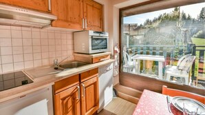 A compact kitchen with wooden cabinets, a microwave, and a dishwasher. A dining area with a table and chairs is visible through a large window. - Ski-in/ski-out & foot of the slopes, Ground floor, swimming pool, terrace, parking, 27m², Les Gets (Les Gets)