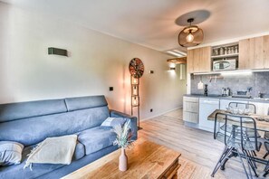 Uma sala de estar moderna com um sofá azul, uma mesa de centro de madeira e uma área de cozinha com micro-ondas e armários.