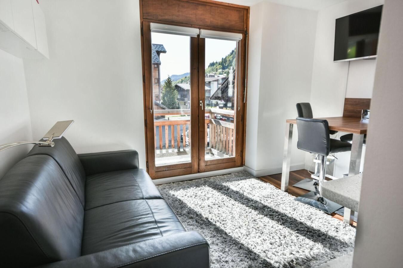 A modern living room with a black leather sofa, a dining area with a table and chairs, a TV mounted on the wall, and a sliding glass door leading to a balcony with a view of buildings and trees.