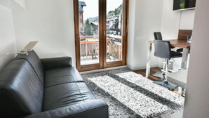 Una sala de estar moderna con un sofá de cuero negro, una zona de comedor con mesa y sillas, un televisor montado en la pared y un ventanal corredizo que da a un balcón con vistas a edificios y árboles.