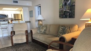A living room with a beige sofa, two wooden chairs, a patterned rug, a kitchen in the background, and a wall-mounted lamp. - Direct Ocean Views, Luxe Living #506 (Cape Canaveral)