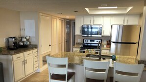 A kitchen with white cabinets, a stainless steel refrigerator, a microwave, and a stove. There are two white chairs with brown cushions at the island. - Direct Ocean Views, Luxe Living #506 (Cape Canaveral)