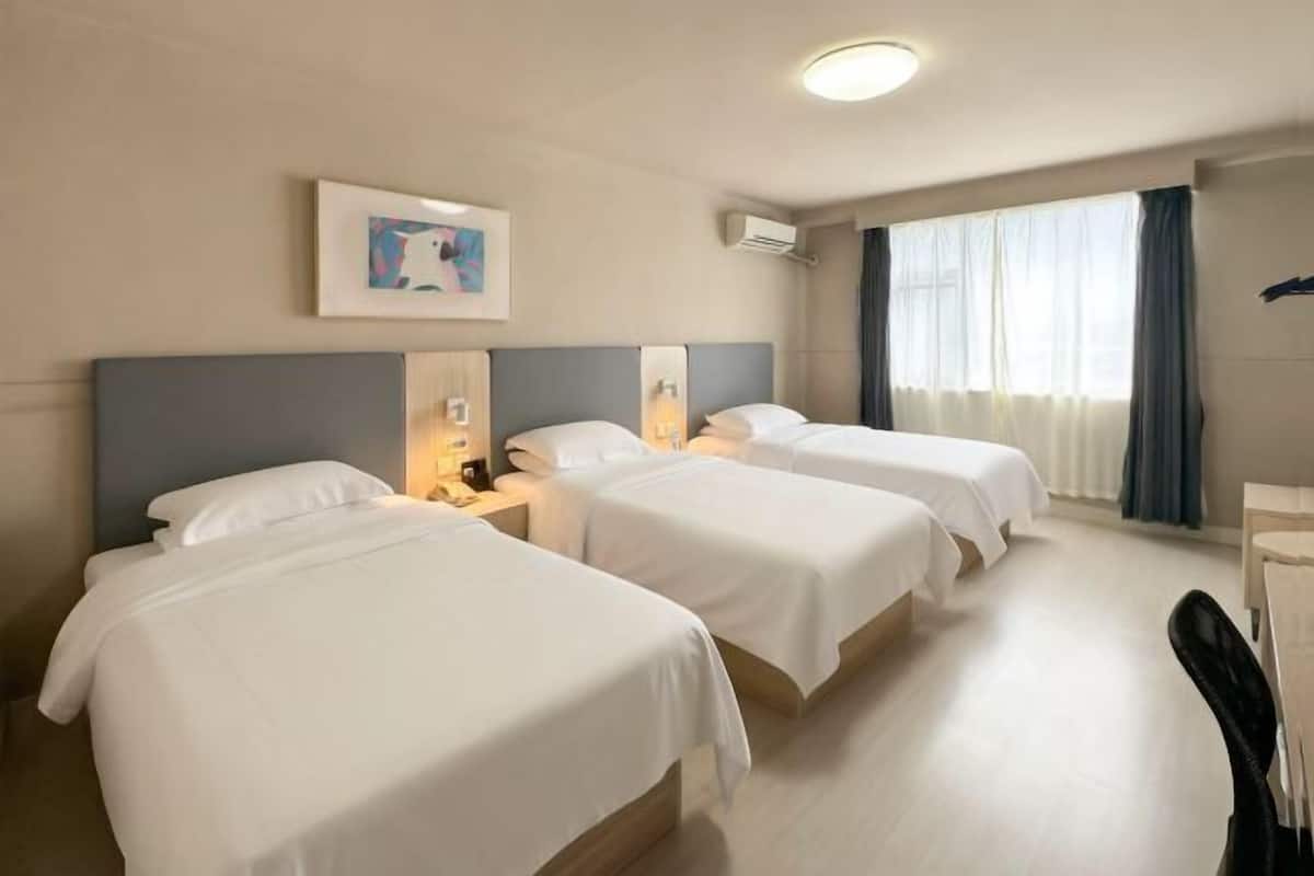 A hotel room with three beds, a desk, a chair, a window with curtains, an air conditioning unit, and a framed picture on the wall.