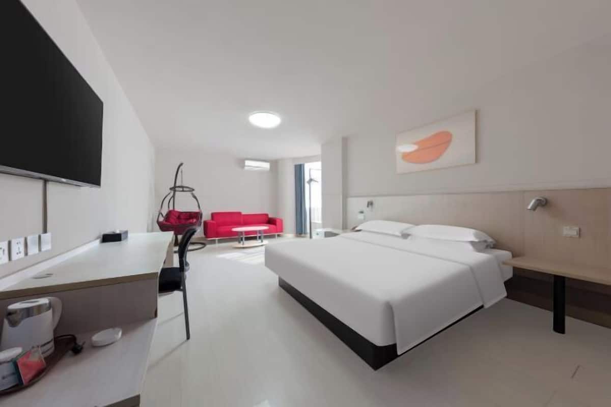 A modern hotel room with a bed, a desk with a coffee maker, a red sofa, and a large window.