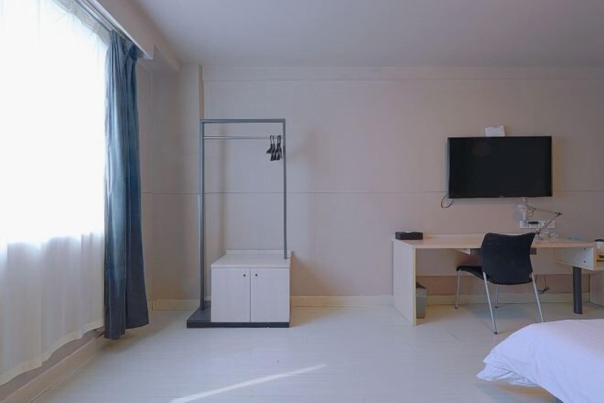 A hotel room with a bed, a desk with a chair, a TV, a wardrobe, and a shower area.