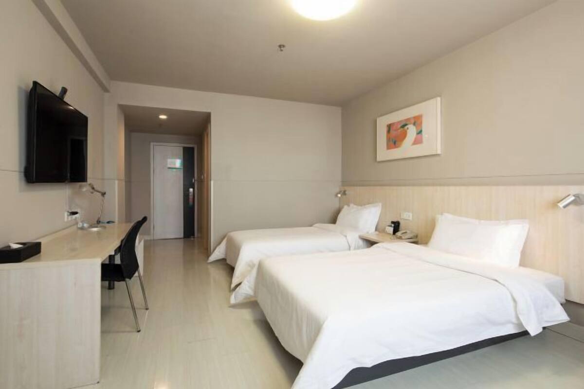 A hotel room with two beds, a desk with a chair, a flat-screen TV mounted on the wall, and a painting above the bed.